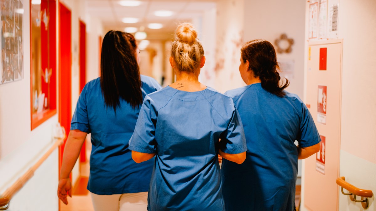 Pflegepersonen gehen in typischer Kleidung einenFlur entlang