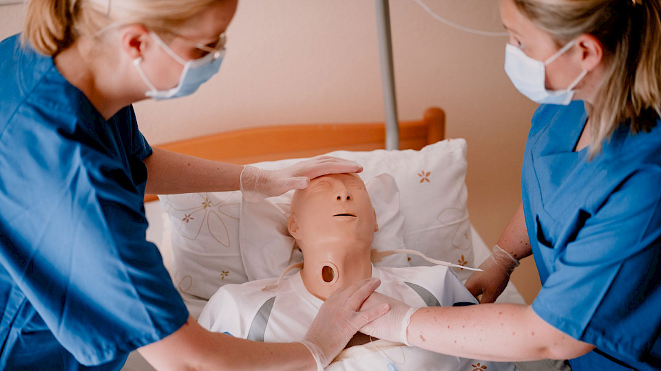 2 Pflegekräfte am Bett, die an einer Schulungspuppe üben