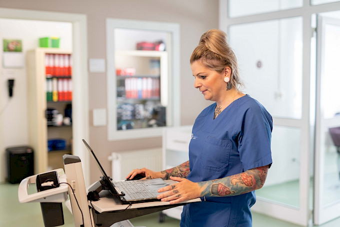 Pflegefachkraft in typischer Kleidung für Intensivpflege arbeitet am Computer. Im Hintergrund ist ein Büro zu sehen.
