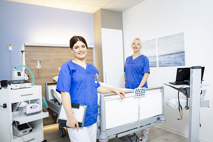 Pflegefachkräfte stehen am Bett eines Patientenzimmers umgeben von Hilfsmitteln der Intensivpflege wie Beatmungsgerät und Absaugpumpe